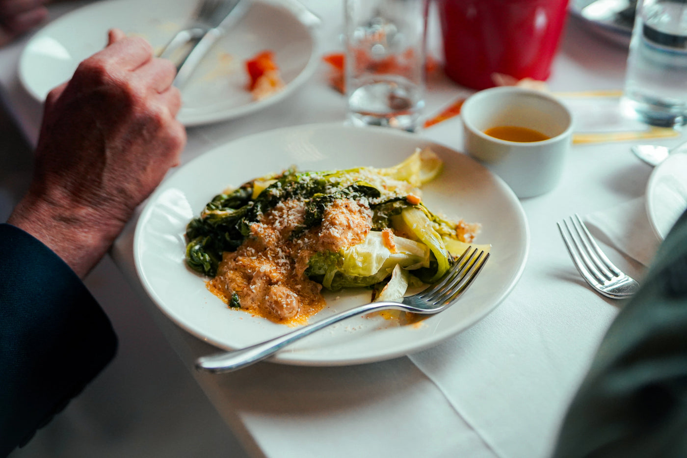 Person holding a fork over a plate of food with greens and sauce on a dining table.
