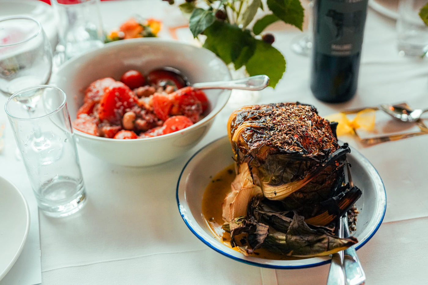 Dish of food on a table with a bowl of fruit and a bottle of wine in the background.