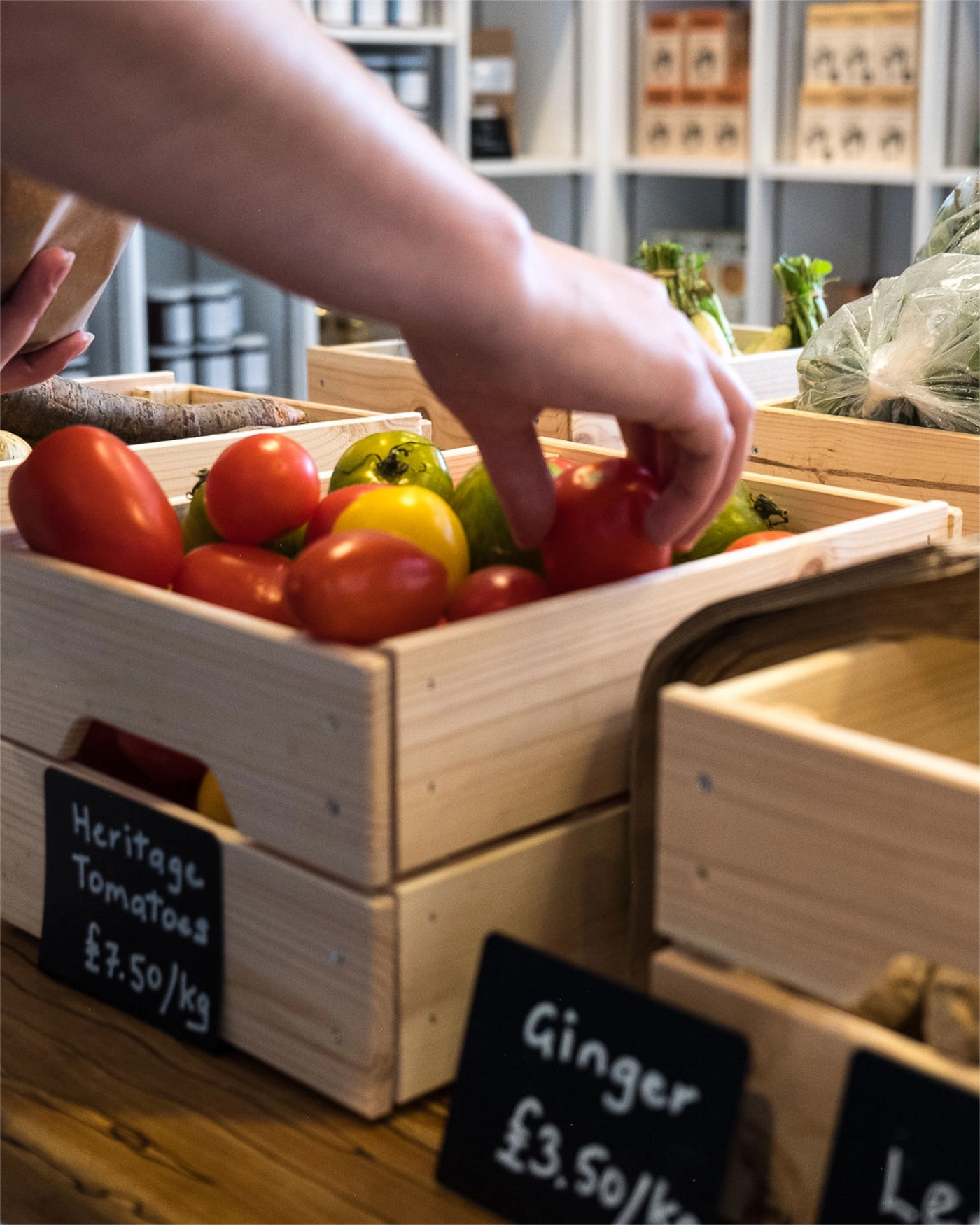 Person selecting tomatoes from a wooden crate with signs indicating prices at a market.