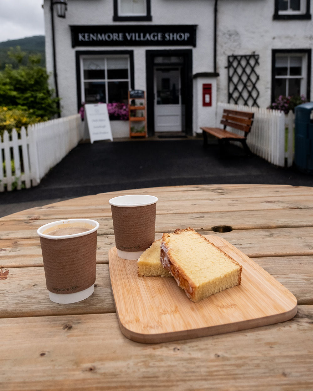 Two paper cups with a sandwich on a wooden board in front of Kenmore Village Shop.