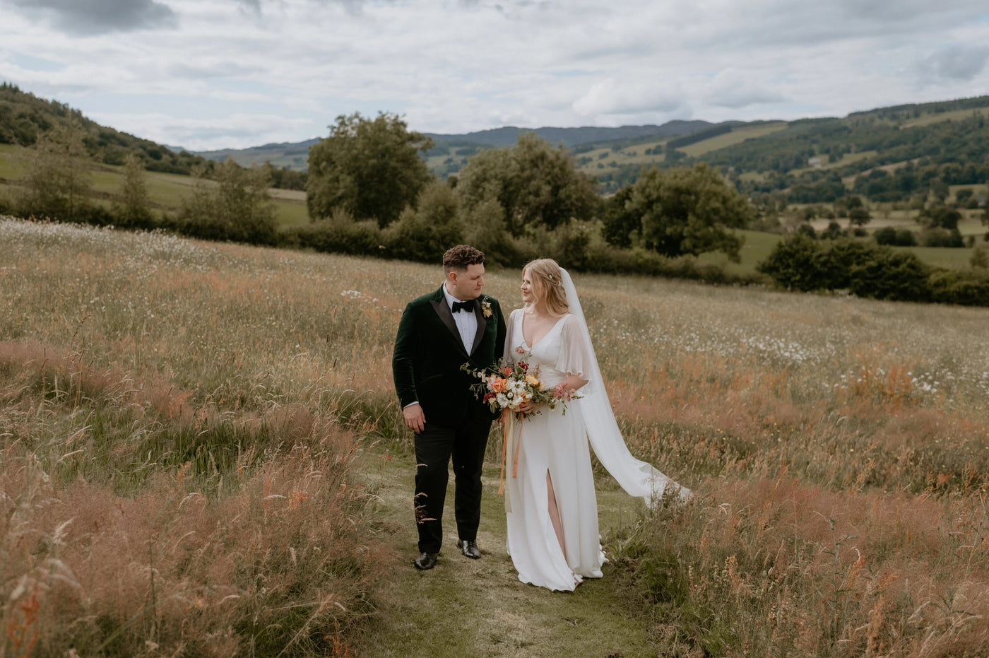Couple in wedding attire standing in a field with mountains in the background