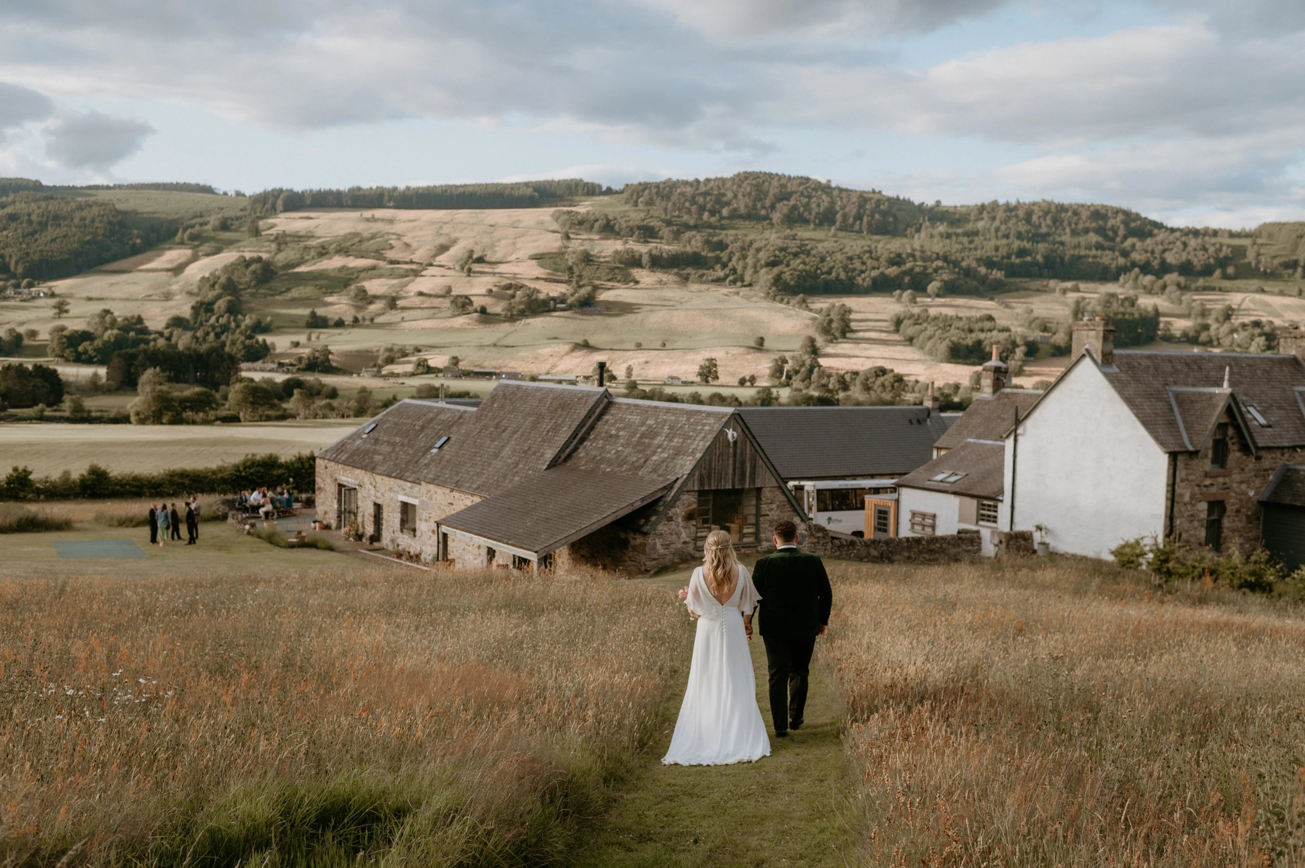 Couple walking through a field with a scenic landscape and buildings in the background