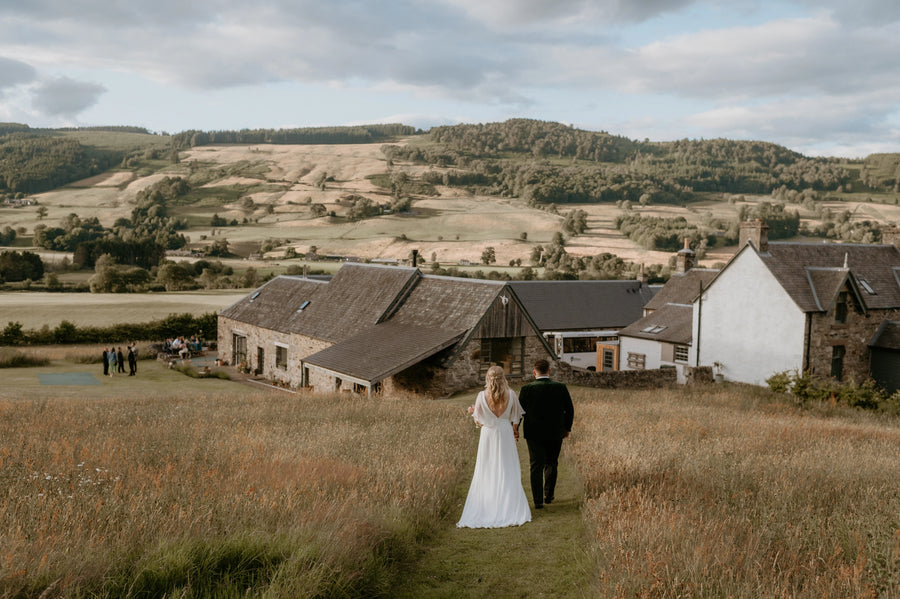 Couple walking through a field with a scenic landscape and buildings in the background