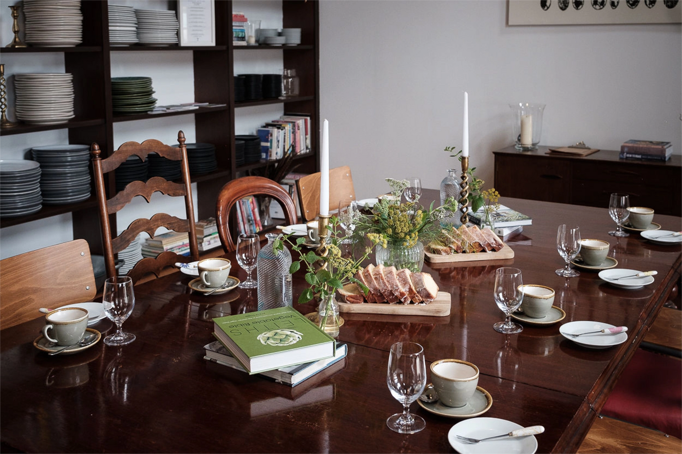 Dining table set for a meal with plates, cups, and cutlery, surrounded by chairs and a bookshelf.