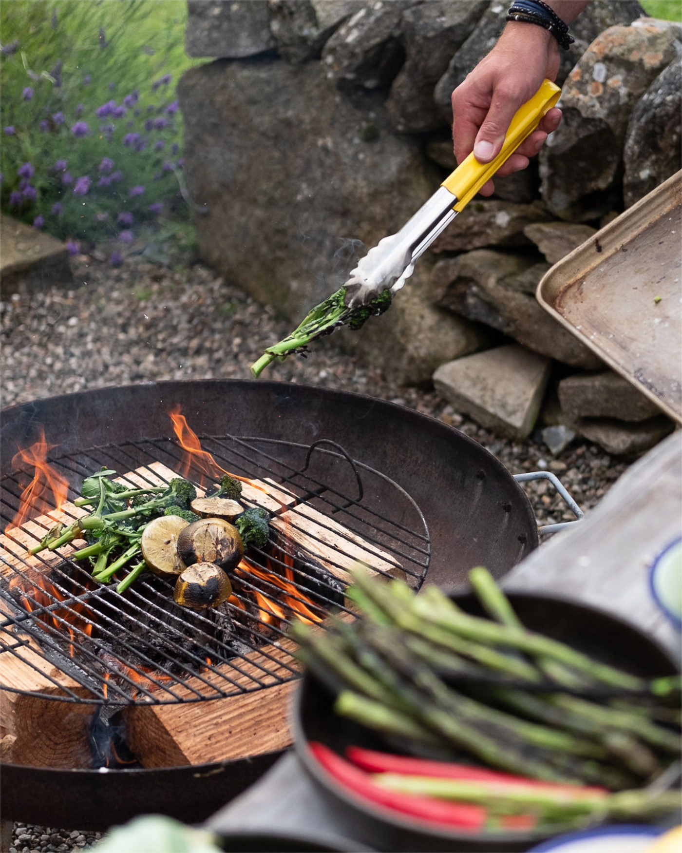 Person grilling vegetables on a barbecue with a stone wall and plants in the background