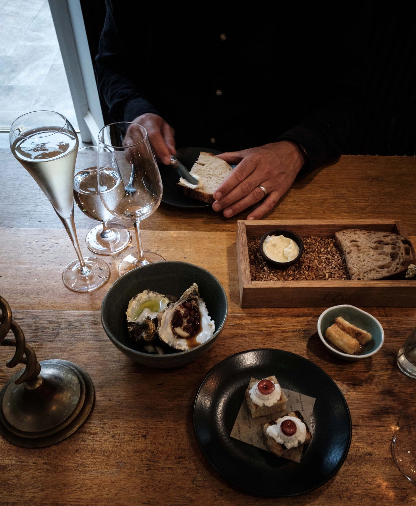 Person at a table with food and drinks, including bread and small dishes on a wooden surface.