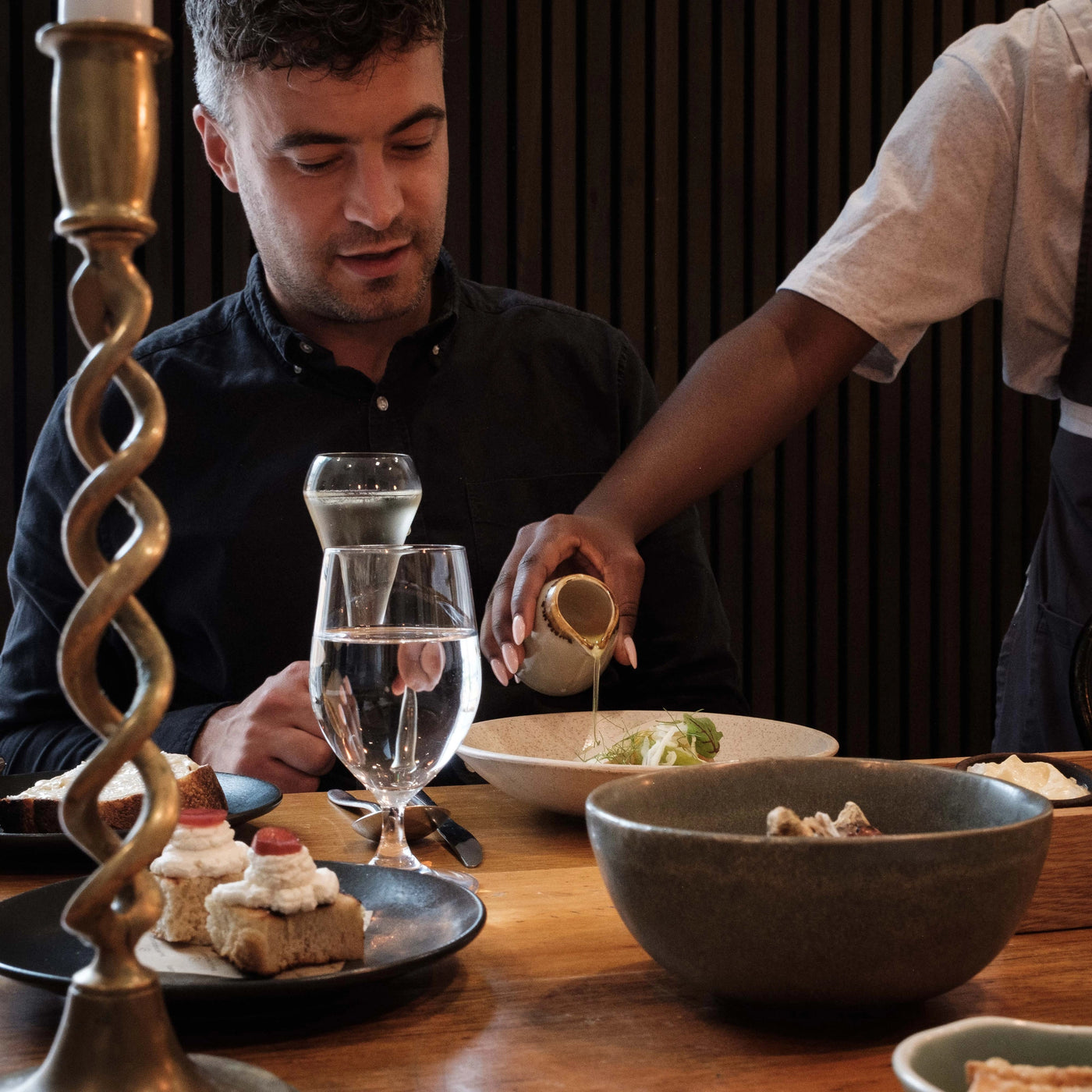Man sitting at a table with a server pouring sauce onto his dish, surrounded by food and drinks.