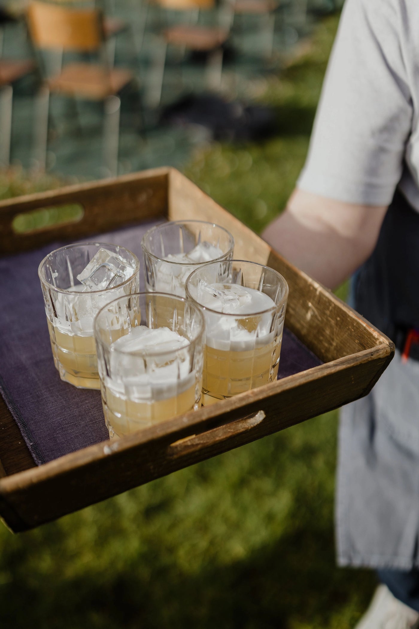 Person holding a tray with four glasses of cocktails and ice cubes outdoors.